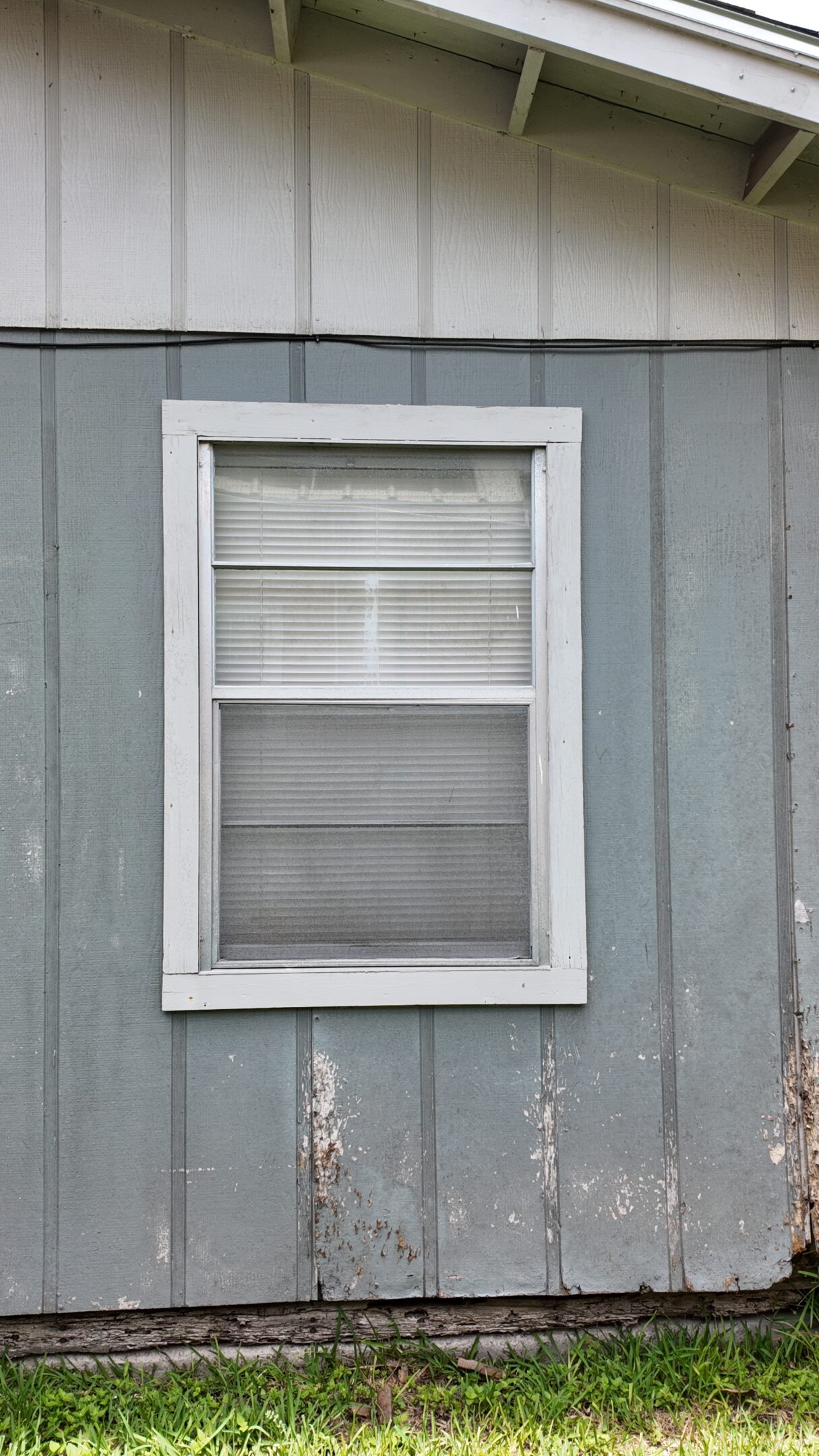A simple white-framed window with closed blinds on a gray wooden wall.