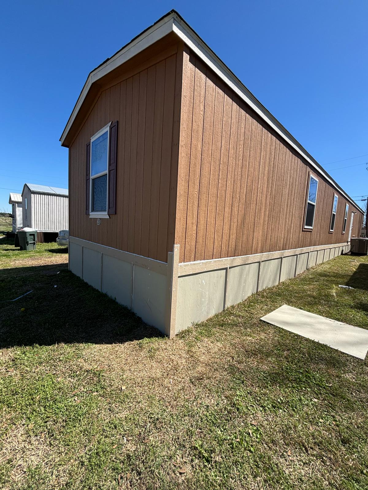 Corner of a wooden mobile home with a concrete foundation under clear blue sky.