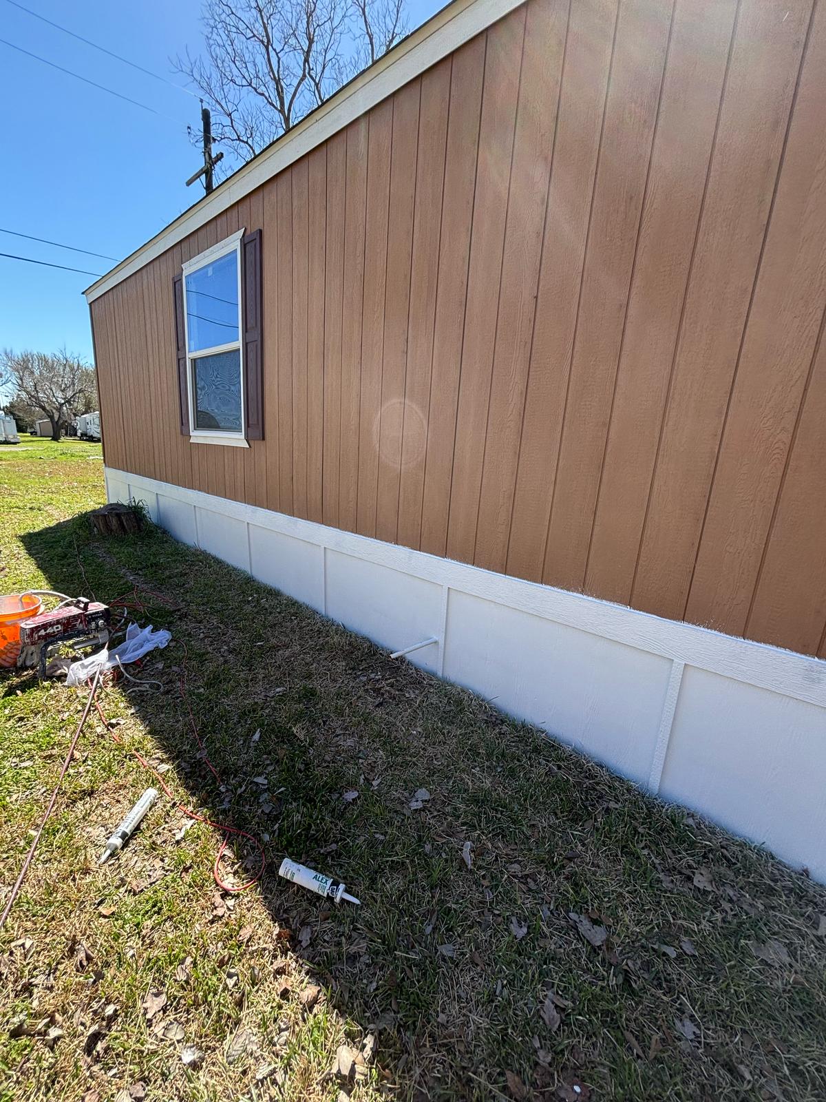 Side view of a brown and white mobile home with grass and some scattered debris nearby.