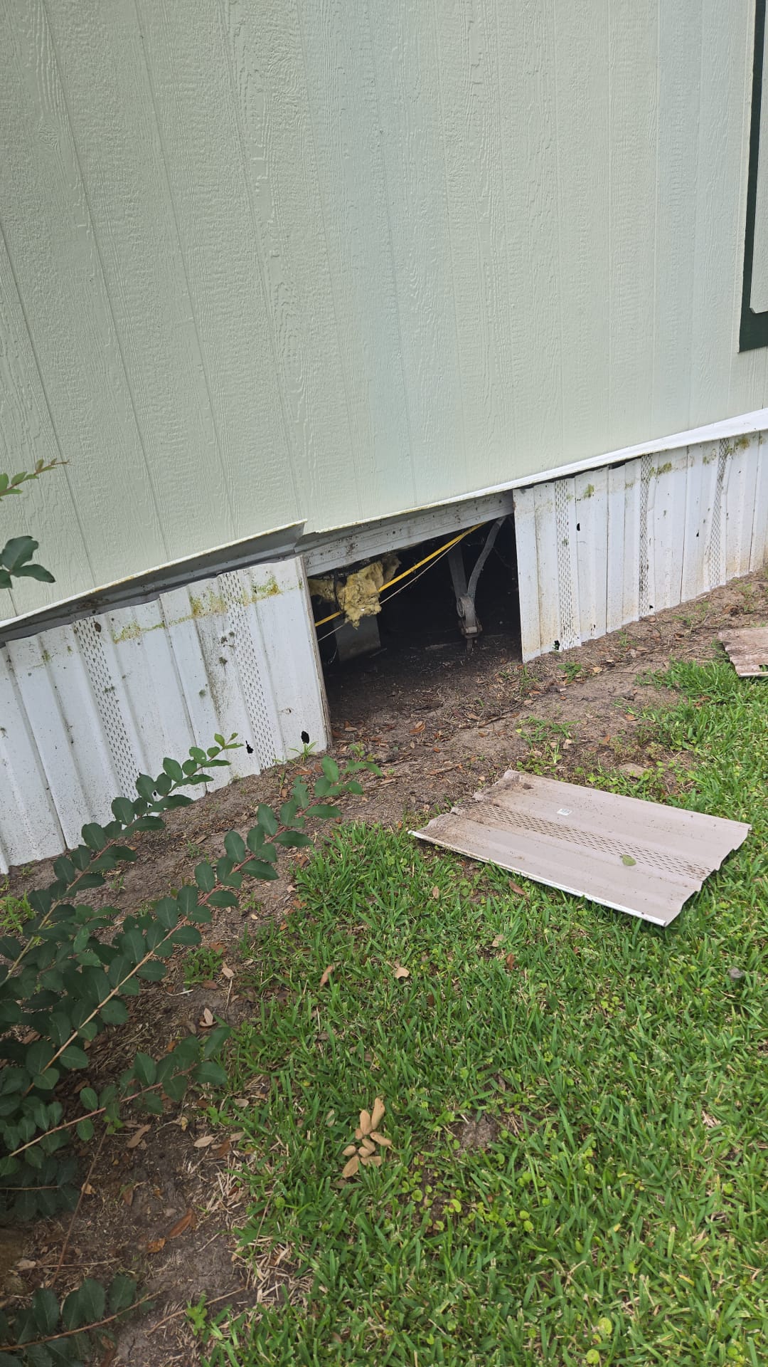 Open crawl space under a house with a loose panel on the grass.