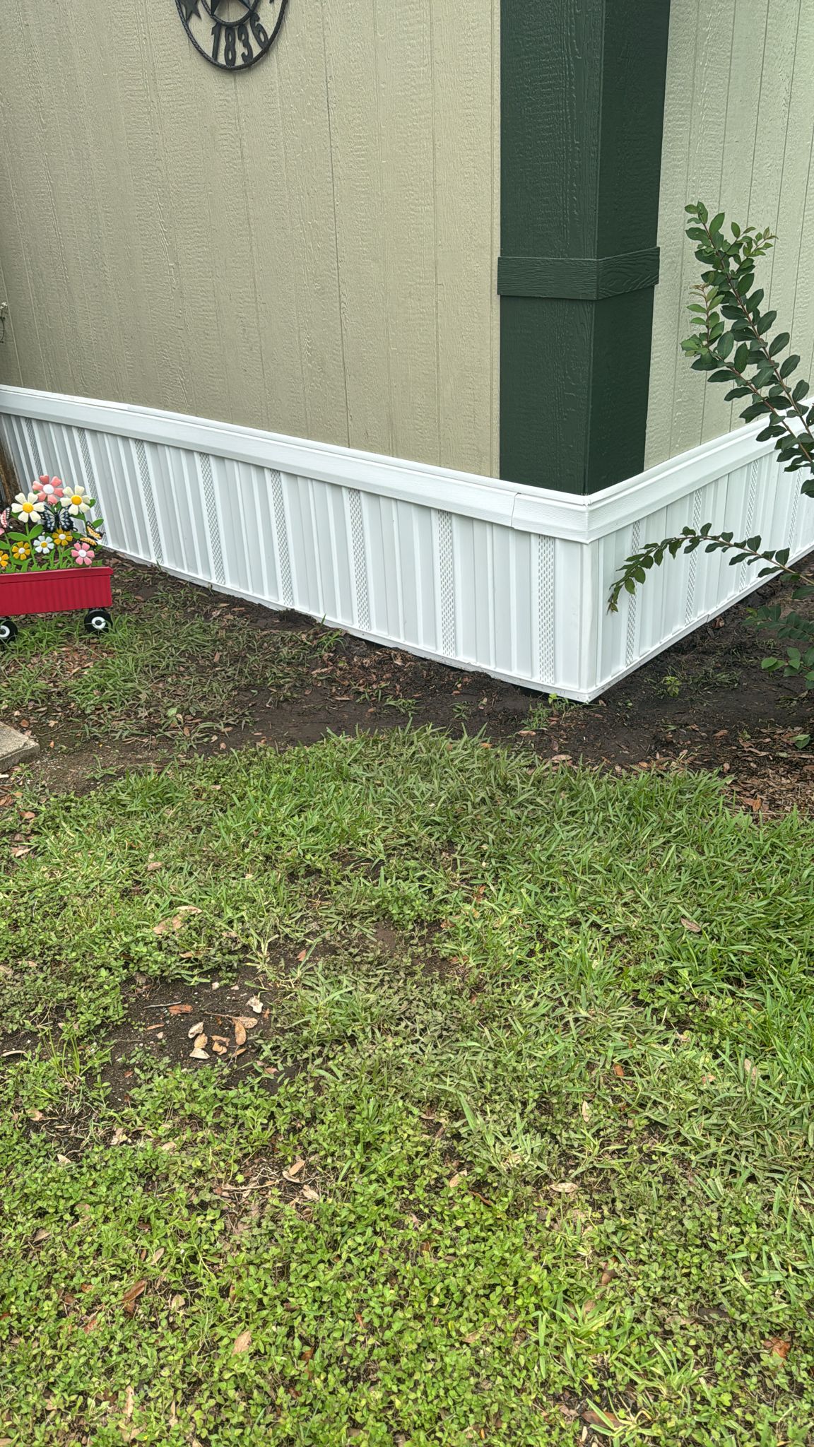A corner of a house with white siding and some grass and soil around it.