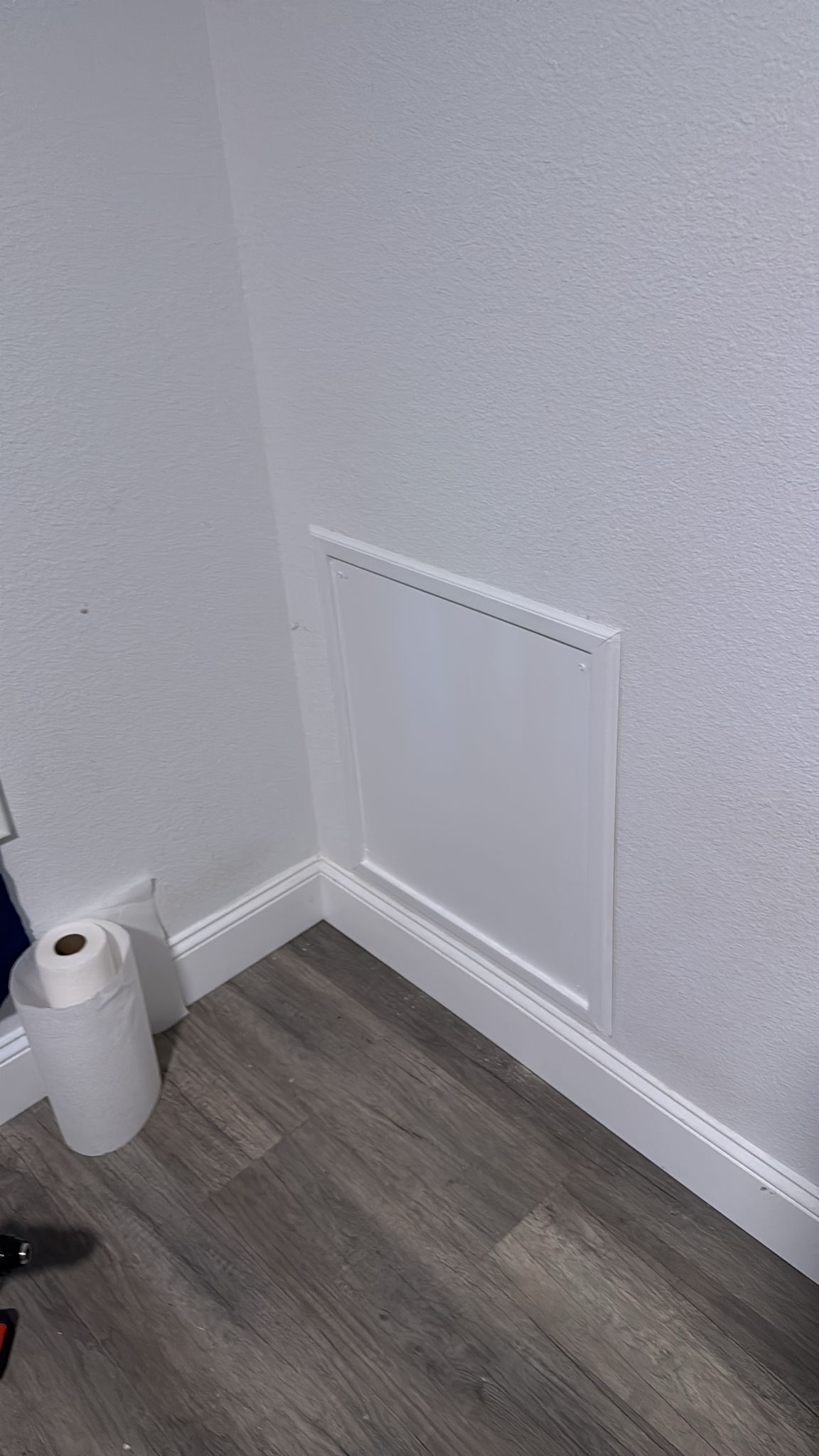 A white wall with a small access panel near the floor and a roll of toilet paper on the floor.