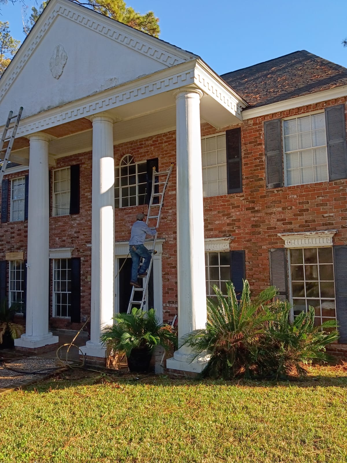 Person on ladder cleaning gutters of a brick house with white pillars.